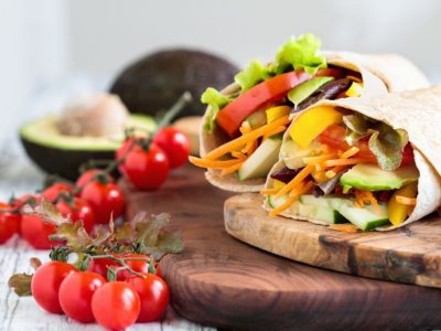A healthy lunch or dinner of a vegan / vegetarian wrap made with  argula lettuce, sliced tomatoes, cucumbers, avocado, bell peppers and carrots. Selective focus on sandwich in front.